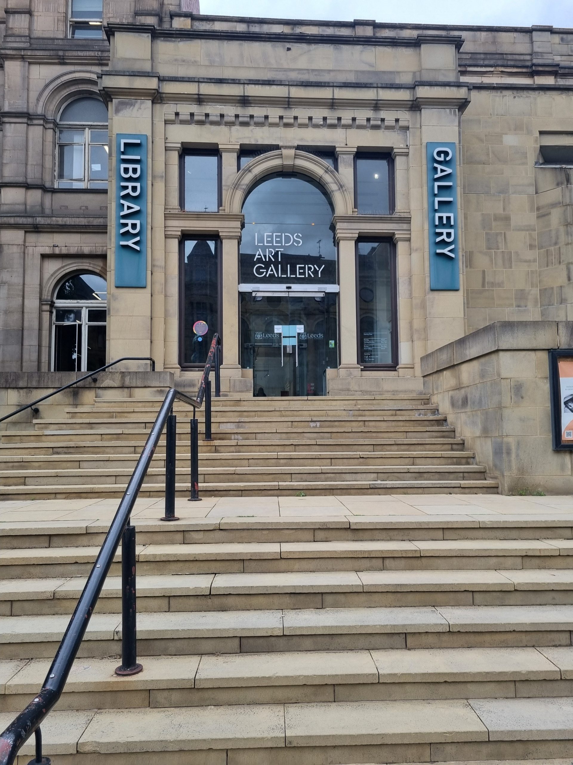 Art Gallery entrance, up a flight of steps with a handrail to the left of the picture. The doors to the gallery are automatic and made of glass. The window above the door says "Leeds Art Gallery". To the right of the door is a sign saying "Gallery" and to the left of the door one saying "Library".