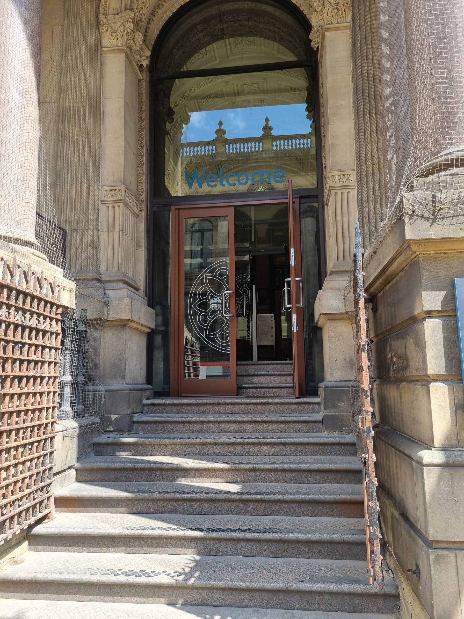 Calverley Street entrance - doors are up a short flight of stairs. There is an open portcullis gate at the bottom of the stairs. The doors into the building are glass and one is propped open. Above the doors is a window, with the word "Welcome" written on it.