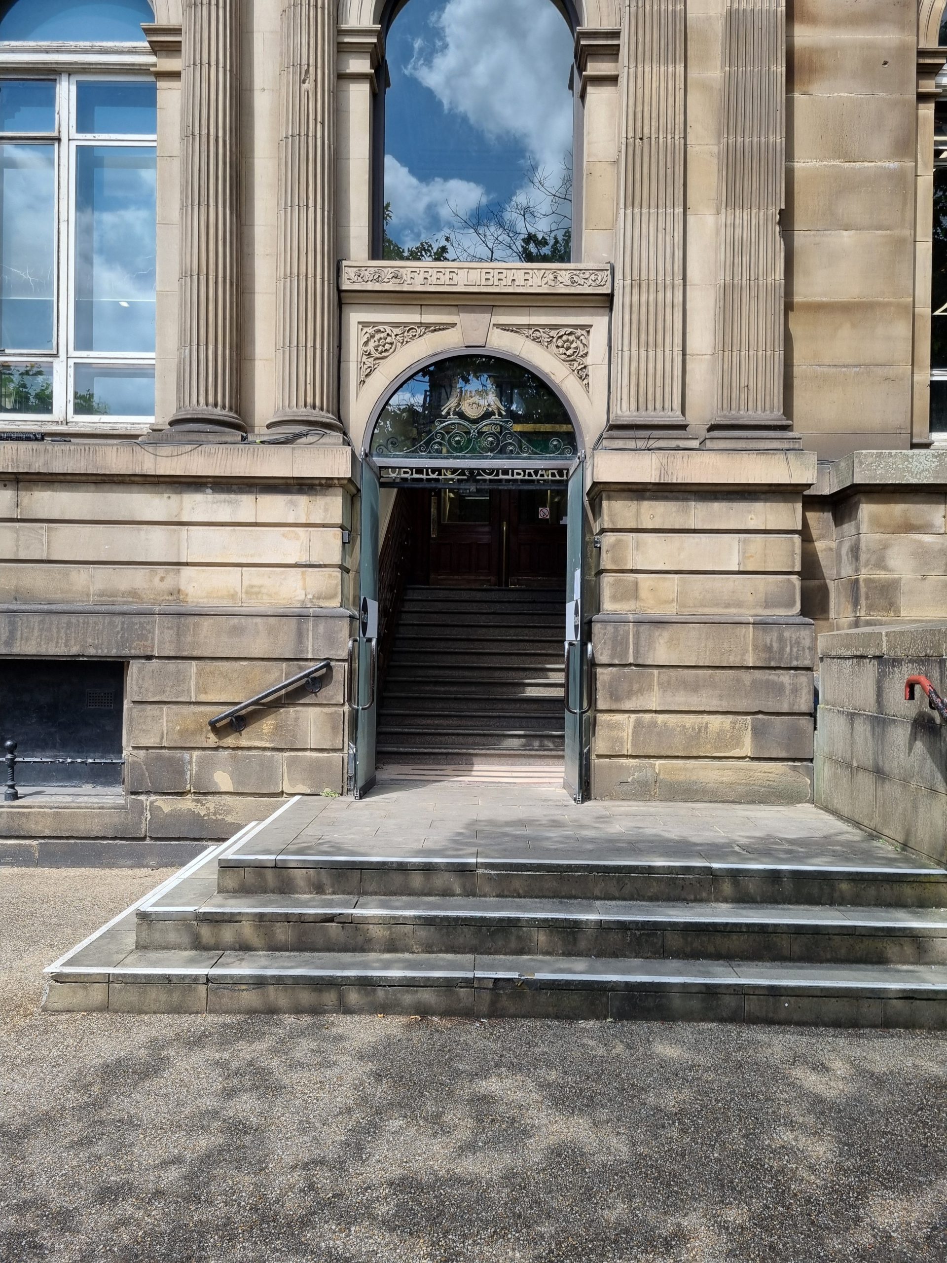 Main entrance to library at ground floor level. Doors are up a set of three steps. Doors are glass and open. Visible above the door is the Leeds coat of arms.